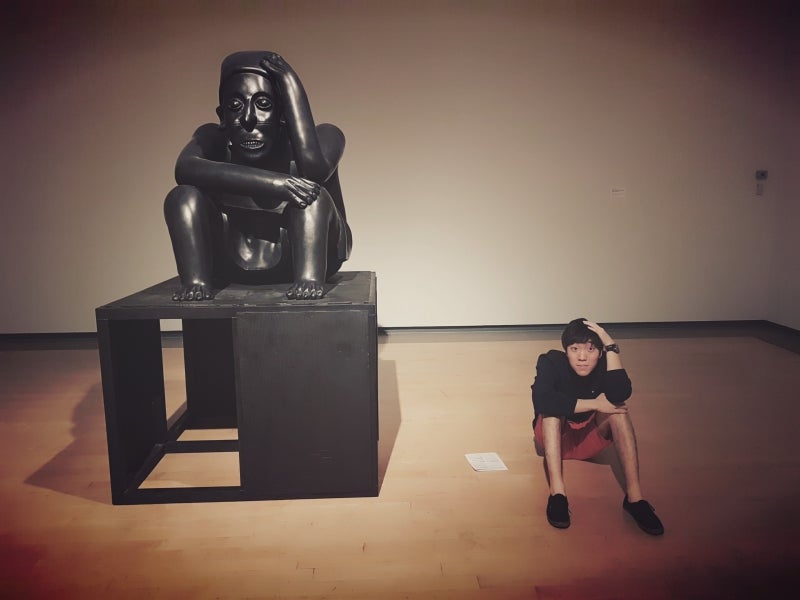 Hojun sitting next a statue imitating it with his hand on his head.