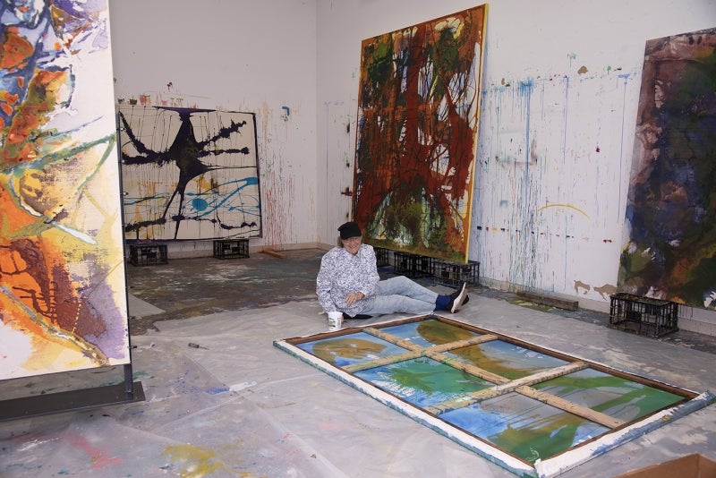 Artist Dona Nelson sits on floor of studio with paintings in foreground and background.