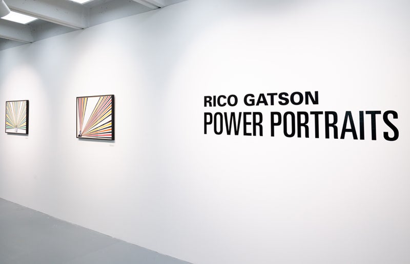White art gallery wall with text reading Rico Gatson: Power Portraits in black lettering, with two framed pieces of art hanging to the left.