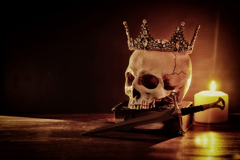 Skull wearing crown placed on top of a closed book, with dagger and candle on a wooden surface. 