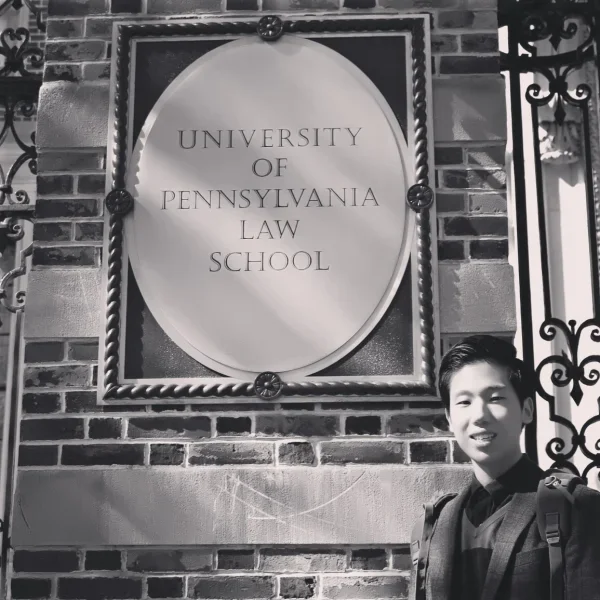 Black and white photo of a person standing next to a University of Pennsylvania Law School sign mounted on a brick wall with decorative ironwork.