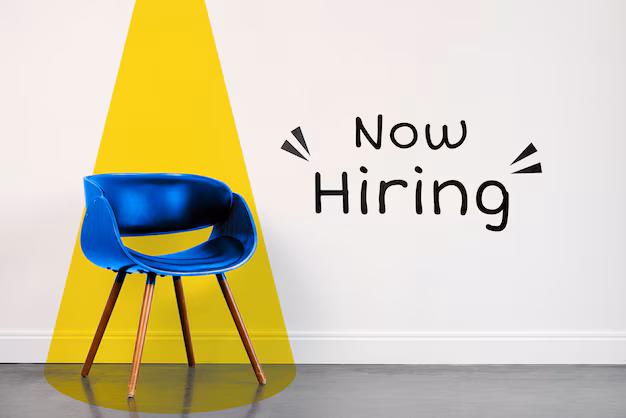 A blue chair illuminated by a yellow spotlight, placed against a white wall with the text 'Now Hiring' written in bold, playful font.
