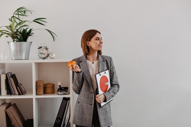 A professional woman in a plaid blazer holding a clipboard with a chart, standing in a modern office setting.