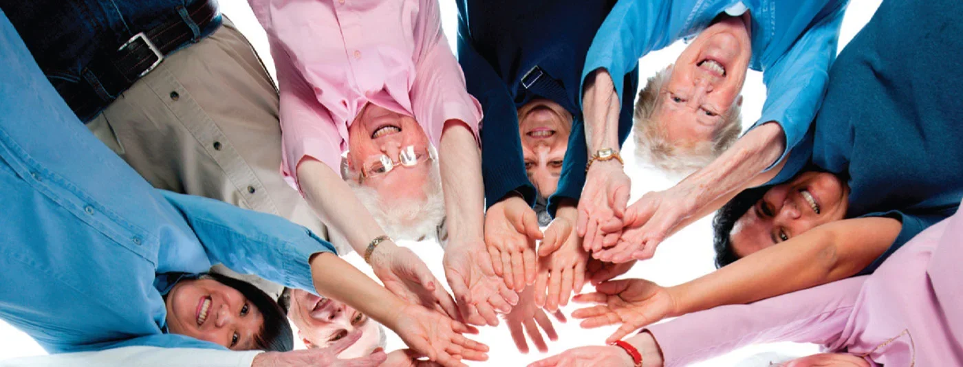 A group of cheerful older adults standing in a circle, reaching into the center with their hands joined together in a symbol of community and togetherness.