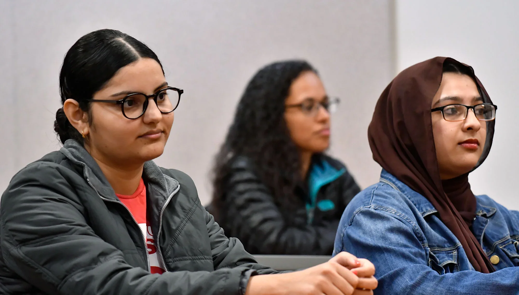 Three students attentively listening in a classroom setting, including two in the foreground wearing glasses and one wearing a headscarf.
