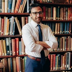 A person with glasses standing in front of bookshelves, wearing a white shirt and tie, with arms crossed.