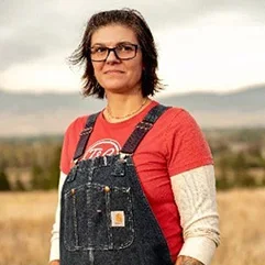 A person with short hair and glasses, wearing a red t-shirt and denim overalls, standing in a field with mountains in the background.