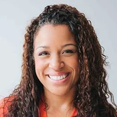 A smiling person with curly hair, wearing an orange shirt, looking directly at the camera.