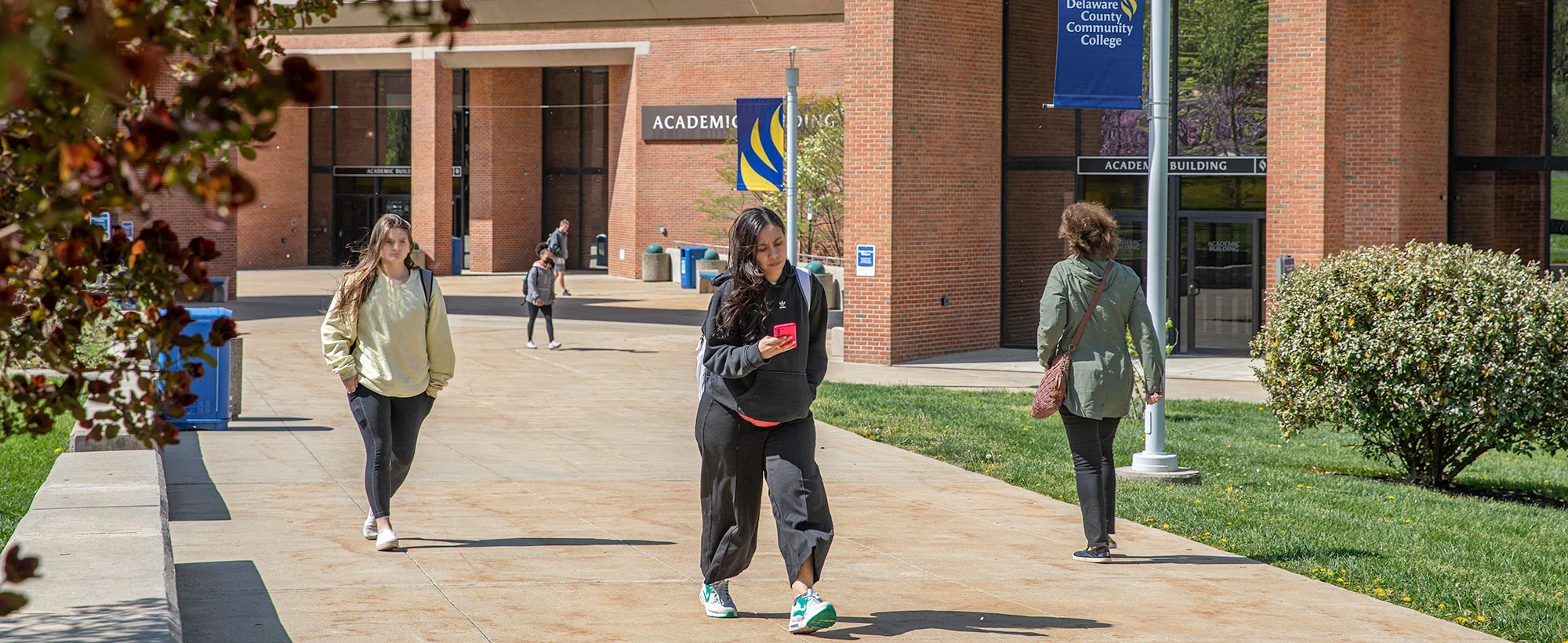Students walking on campus near the Academic Building at Delaware County Community College, surrounded by greenery and campus banners on a sunny day.