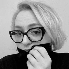 A black-and-white portrait of a person with short blonde hair and glasses, holding their sweater collar close to their face.