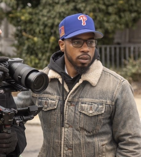 A man wearing a denim jacket, glasses, and a blue baseball cap, standing outdoors next to a professional camera.