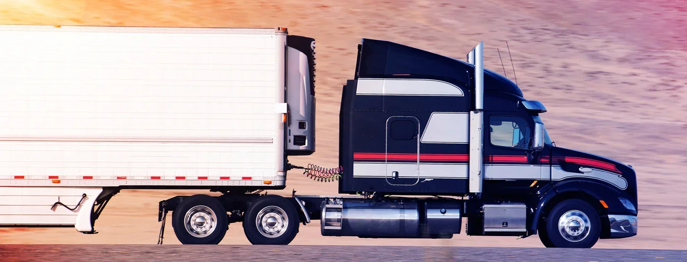 Side view of a black and white semi-truck with a trailer driving on a highway during sunset.