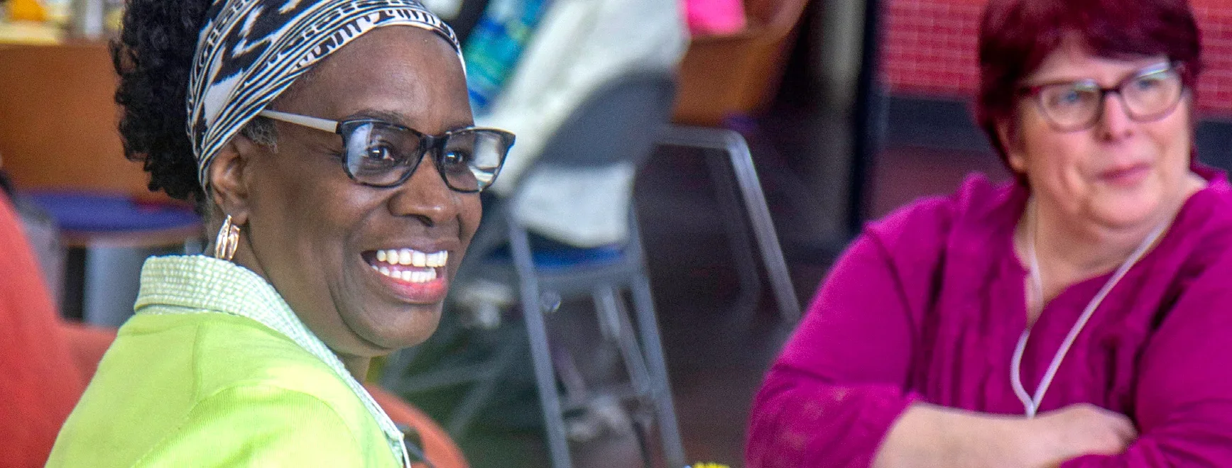 A cheerful woman wearing glasses and a headband smiling during a community event, with another attendee in the background.