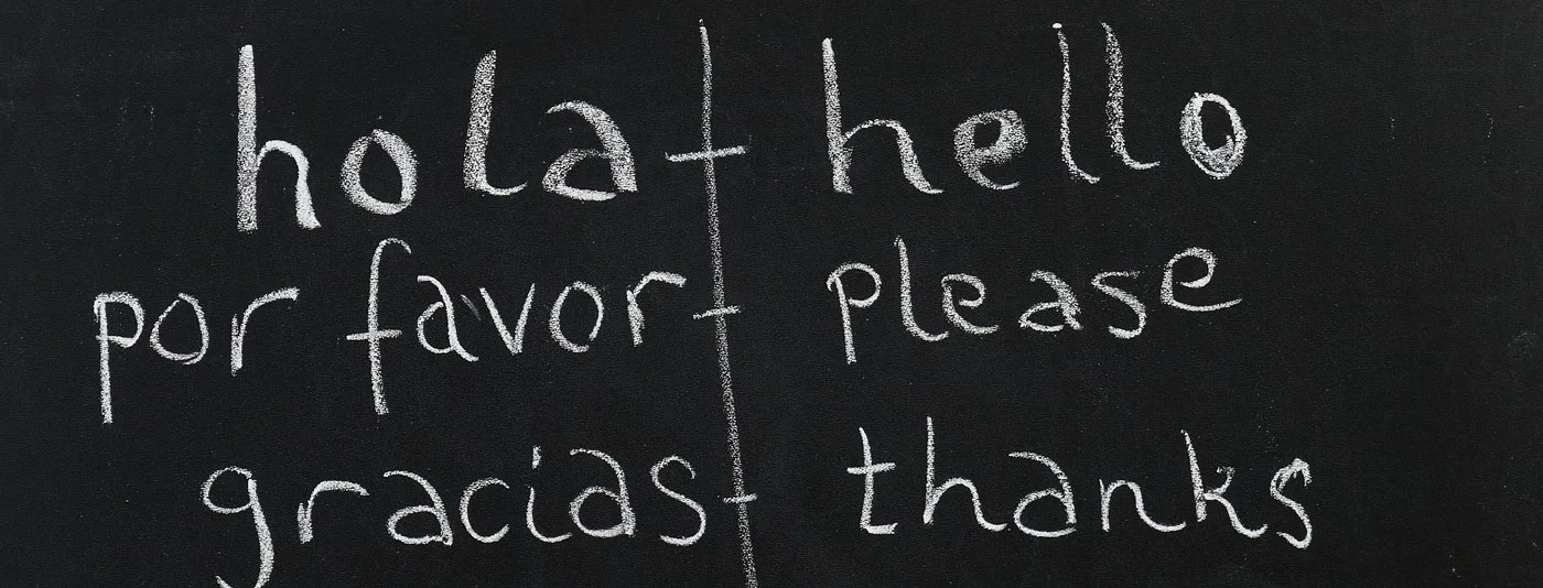 A chalkboard displaying common Spanish words like 'hola,' 'por favor,' and 'gracias' with their English translations 'hello,' 'please,' and 'thanks.
