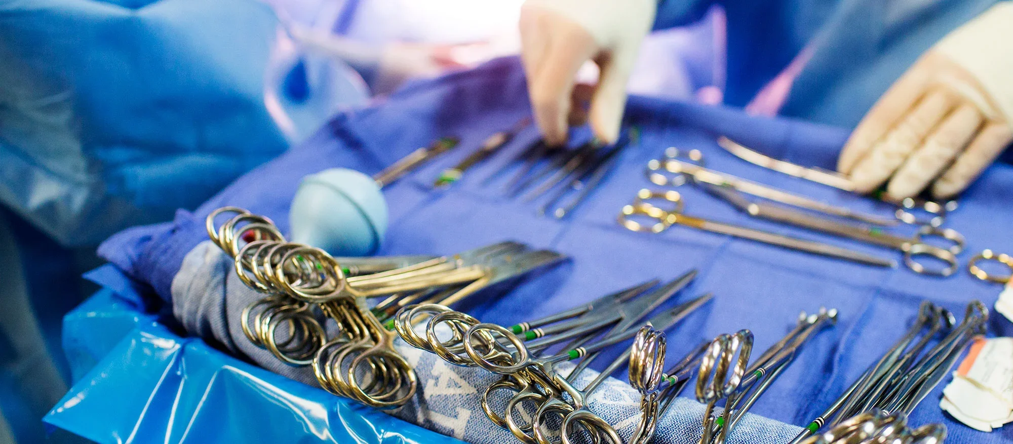 A close-up of surgical instruments arranged on a sterile blue drape in an operating room, with a gloved hand organizing tools in the background.