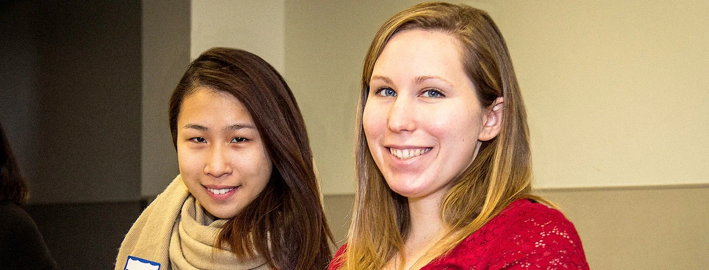 Two smiling students pose together indoors, one wearing a beige scarf and the other in a red lace top. Their expressions convey warmth and friendship, highlighting a diverse and inclusive environment.
