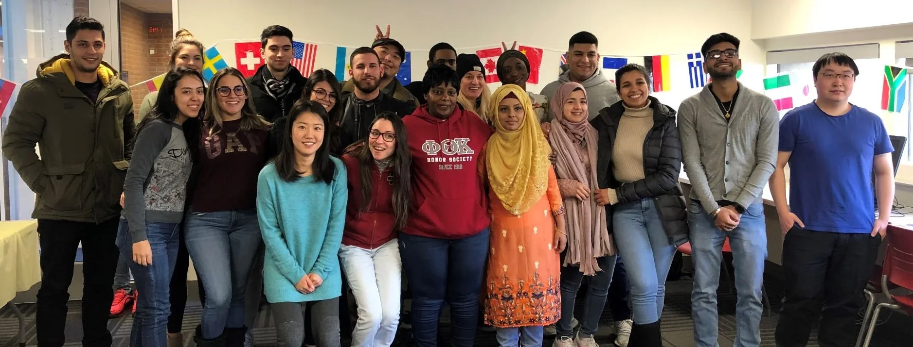 A diverse group of students poses together in a brightly lit space, smiling for the camera. The background features colorful international flags, suggesting an event celebrating global diversity or international students.