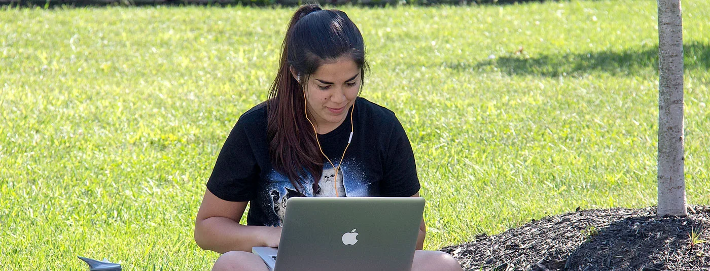 Student sitting on grass outdoors, using a laptop with headphones in a relaxed setting.