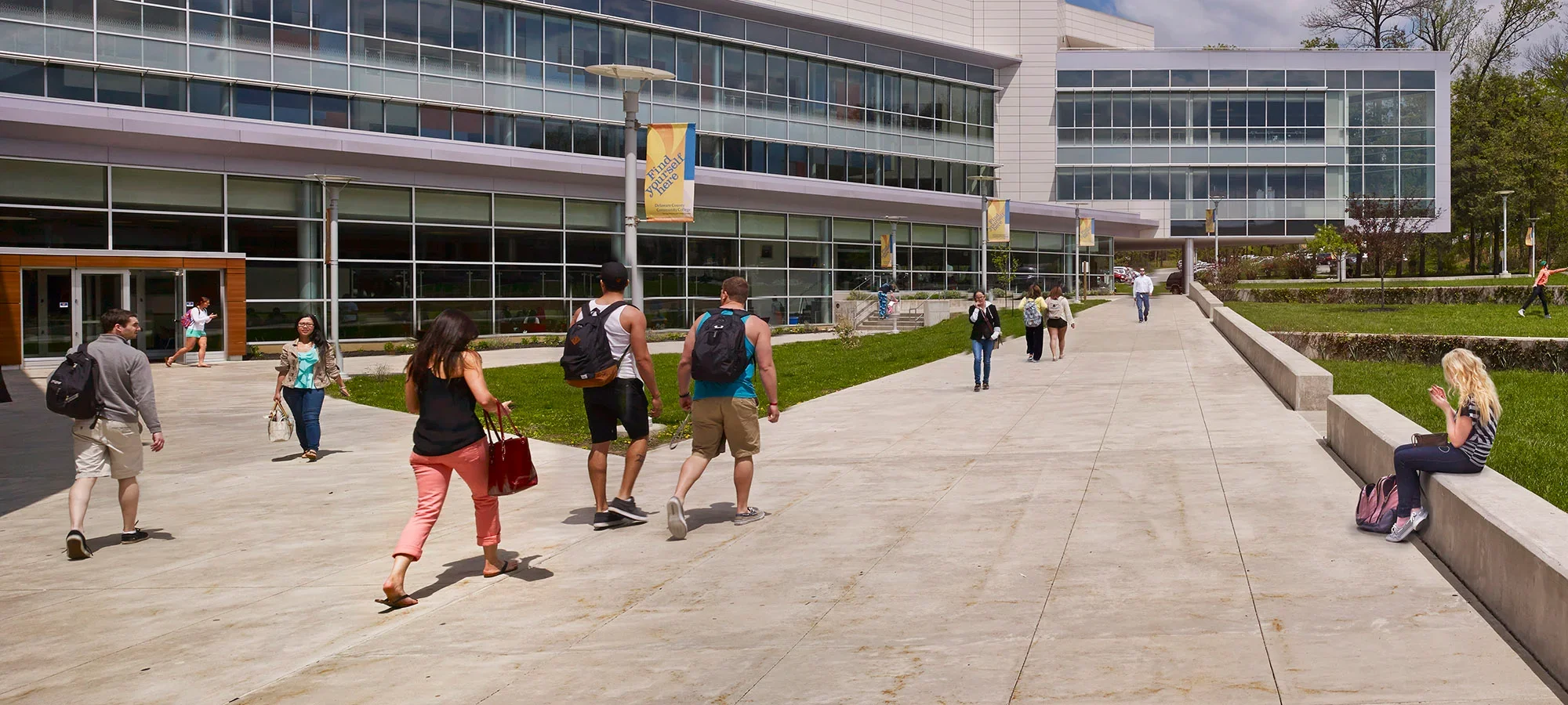 Students walking and sitting outside a modern campus building on a sunny day, with banners displaying motivational messages and a green landscape surrounding the pathway.