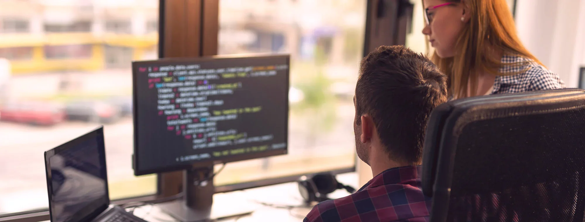 Two programmers working on a project; a man seated at a desk reviewing code on a monitor, with a woman standing beside him, offering guidance