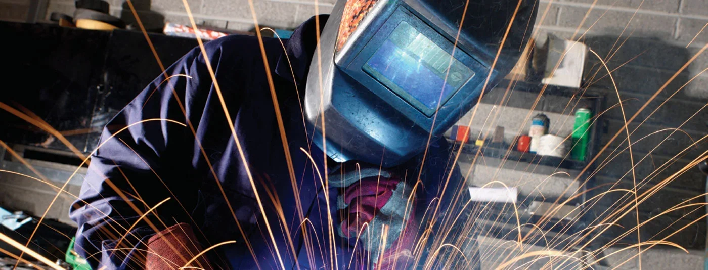 A welder wearing a protective helmet and gear working with metal, emitting bright sparks in a workshop environment.