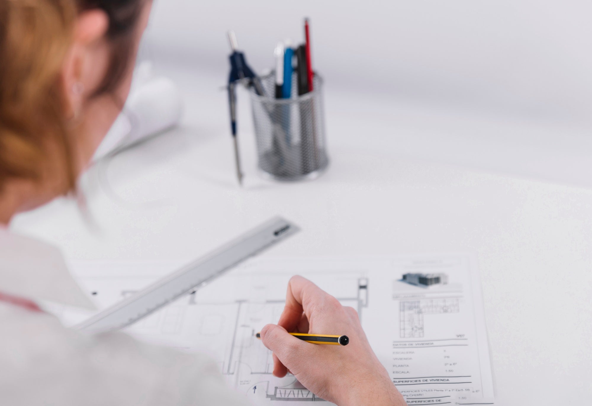 A person holding a pencil and working on architectural or technical drawings at a white desk, with a ruler and a pen holder in the background.