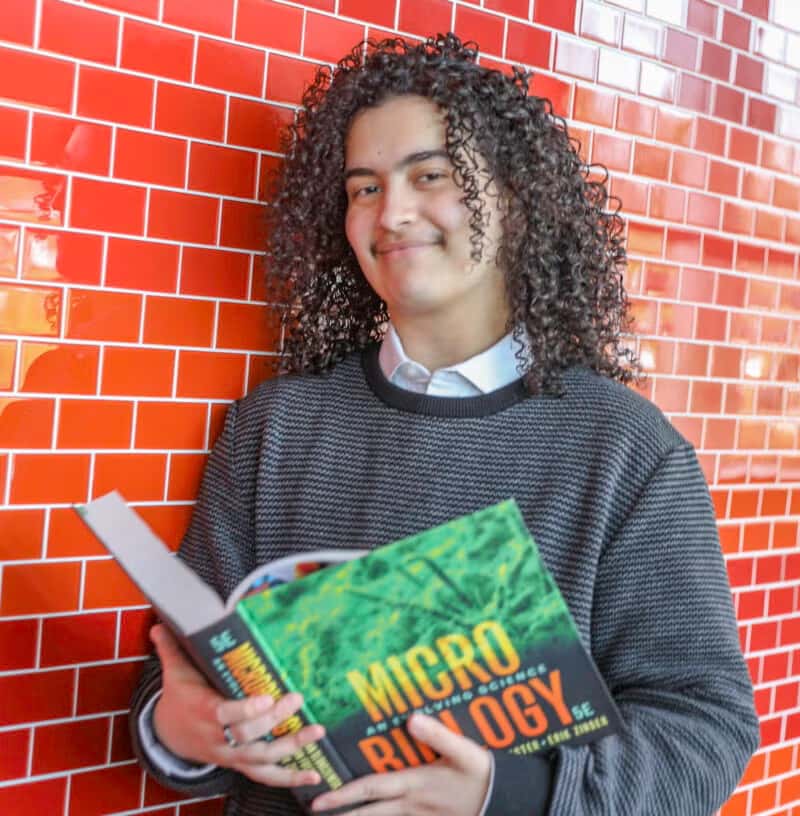 Student with curly hair smiling while holding an open microbiology textbook, standing in front of a red tiled wall.