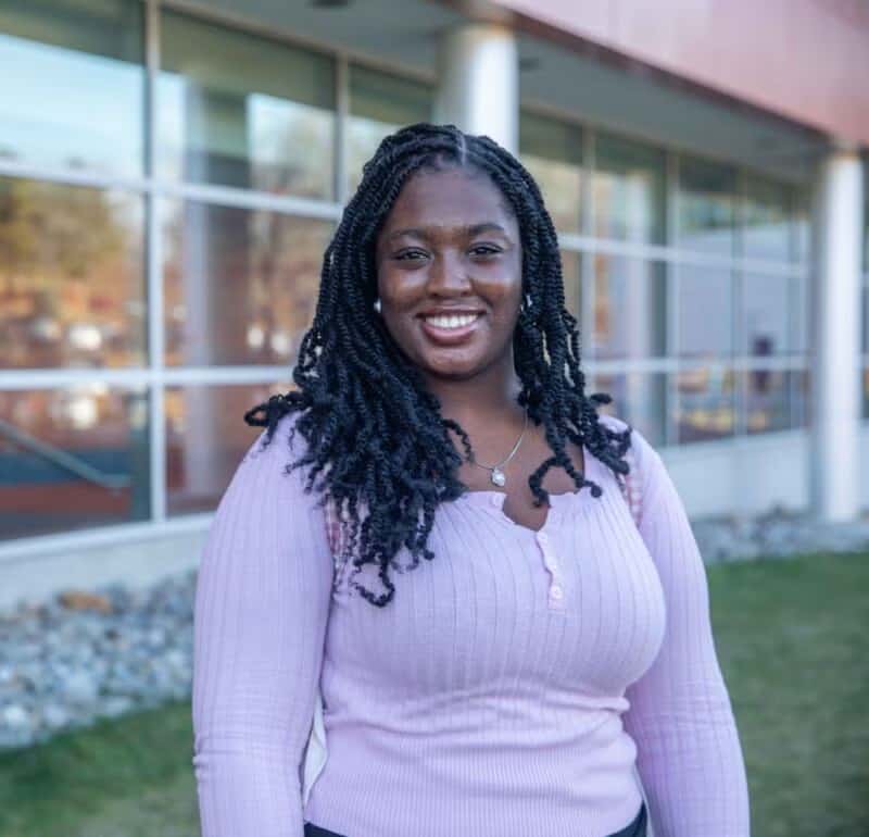 Woman standing outside a modern building with large windows, smiling and wearing a light purple top.