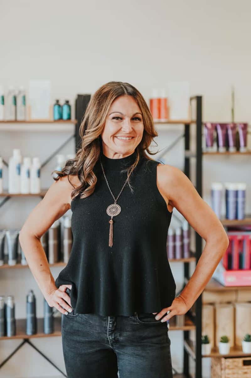 Woman smiling and posing confidently in a salon or beauty store, standing in front of shelves with hair products.