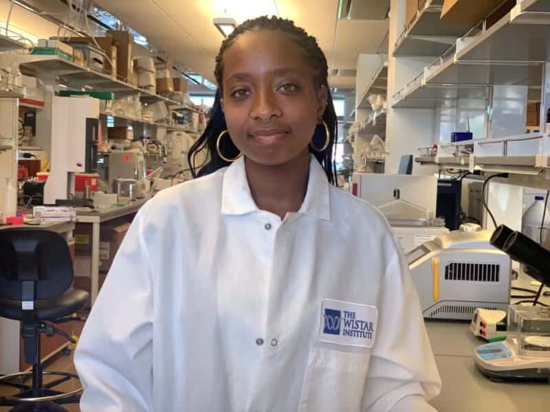 Woman in a lab coat standing in a research laboratory, wearing hoop earrings and looking at the camera, with lab equipment and shelves in the background.
