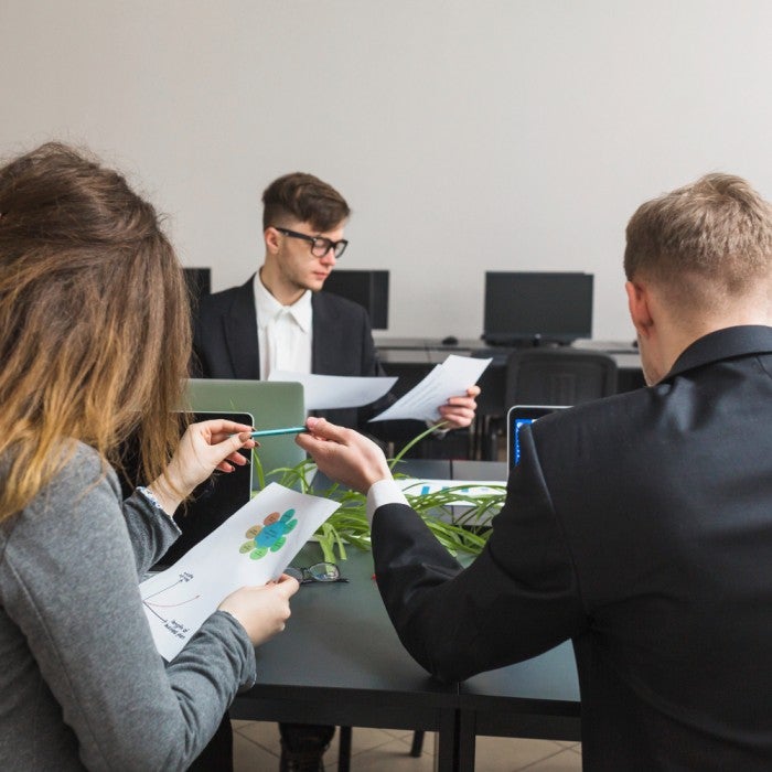 Three individuals in formal attire working together in a modern office setting, reviewing documents and discussing data, with computers in the background.