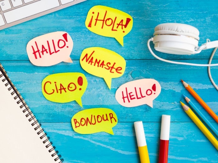 Colorful speech bubbles with greetings in multiple languages, including 'Hallo,' 'Hola,' 'Namaste,' 'Ciao,' 'Bonjour,' and 'Hello,' placed on a bright blue wooden table with headphones, pencils, markers, and a notebook nearby.