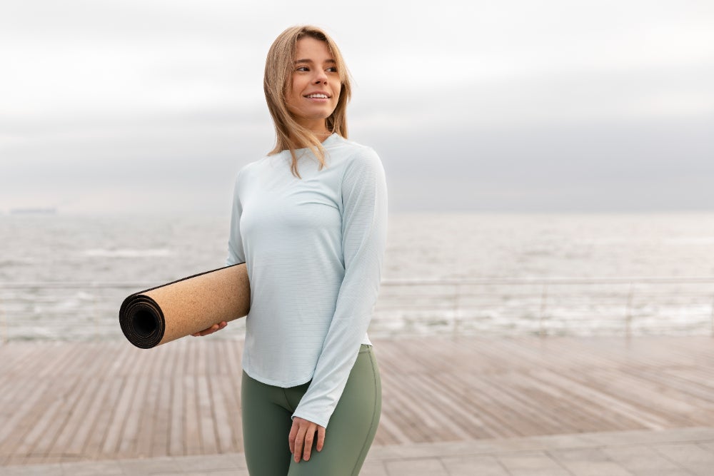 Smiling woman holding a yoga mat while standing on a wooden deck near the sea, dressed in casual workout attire.
