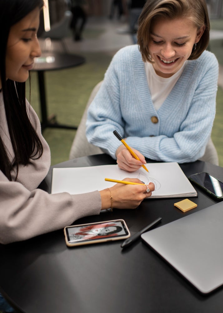 Two women collaborating at a table, one drawing in a notebook while the other observes, with a smartphone, laptop, and stationery on the table.