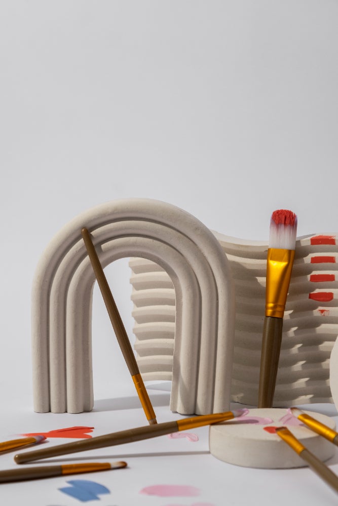 Abstract artistic setup featuring beige ceramic arches and waves, surrounded by paintbrushes and colorful paint strokes on a white surface."