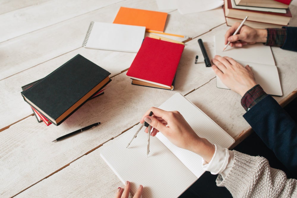 Two people working at a wooden table with notebooks, pens, and a compass, surrounded by stacks of colorful books and stationery.