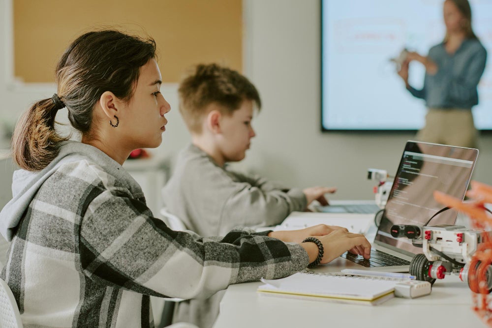 A young student working on a laptop in a classroom, focusing on a coding or robotics project. Another student is seated in the background, and a teacher is visible near the whiteboard in the distance.